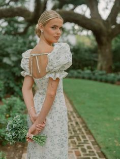 a woman in a white dress is holding flowers and looking off to the side while standing on a brick path