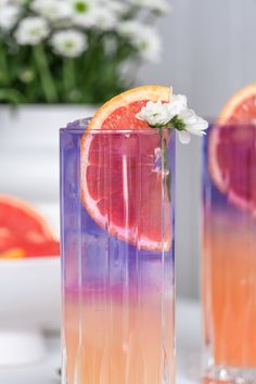 two glasses filled with orange slices and flowers on top of a white table next to watermelon slices