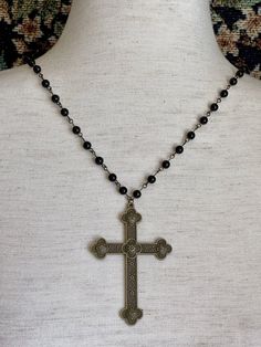 Step back in time with this intricate hand-assembled metal cross rosary-style necklace, featuring a long black beaded chain, and quatrefoil design bronze colored cross pendant. Perfect for tying together a Gothic Medieval, or perhaps Victorian Mourning styled look. This design is part of our handmade collection, making each piece unique. Sizing: One SizeChain Length - 30 inches / 76 CentimetersPendant Length - 3 inches / 7.6 CentimetersTotal Length Laid Flat - 18 inches / 45.5 Centimeters Care I Black Metal Necklace With Antique Finish, Medieval Style Black Metal Necklace, Black Spiritual Jewelry With Antique Finish, Handmade Adjustable Black Cross Necklace, Handmade Black Cross Necklace, Handmade Medieval Black Necklace, Medieval Black Metal Necklace, Handmade Black Cross Pendant Necklace, Bohemian Black Cross Jewelry