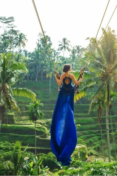 a woman in blue dress swinging on rope with palm trees and rice terraces behind her