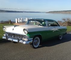 an old green and white car is parked on the side of the road by the water