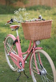 a pink bicycle with a basket full of flowers on the front and back wheel, parked in grass