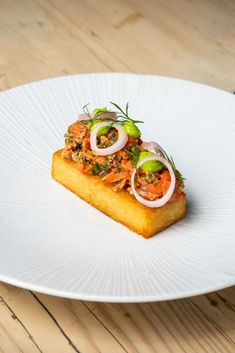 a white plate topped with a piece of cake covered in toppings on top of a wooden table