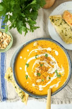 a bowl of carrot soup on a table with bread, nuts and green parsley