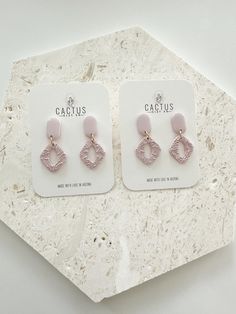 two pairs of pink and gold earrings sitting on top of a white marble table next to a hexagonal object