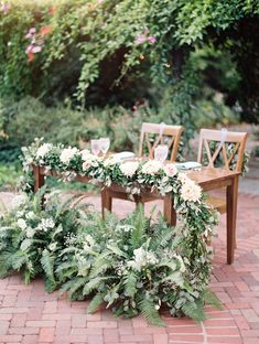 an outdoor table with flowers and greenery on it, surrounded by wooden dining chairs