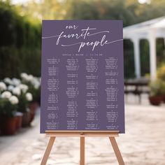 a purple and white wedding seating plan on an easel in front of some potted plants