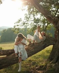 three women in white dresses sitting on a tree branch with the sun shining behind them