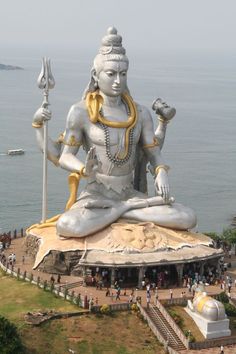 a large statue sitting on top of a lush green hillside next to the ocean with people standing around it