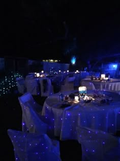 tables and chairs covered in white cloths with blue lights on them are lit up at night