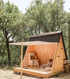 a small wooden cabin in the woods with a woman sitting on it's bed