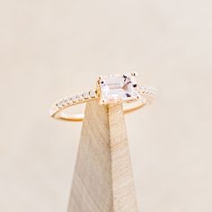 an engagement ring with a square cut diamond on the side, sitting on top of a wooden stand