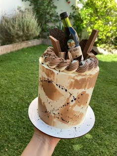 a hand holding a cake with chocolate frosting and wine bottle on top in the grass