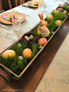 an easter table setting with grass and painted eggs in a long metal tray on top of the table