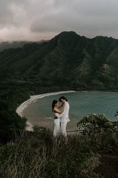 two people standing on top of a hill next to the ocean and hills in the background