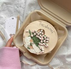 a cake with flowers on it sitting in a box next to a person's hand