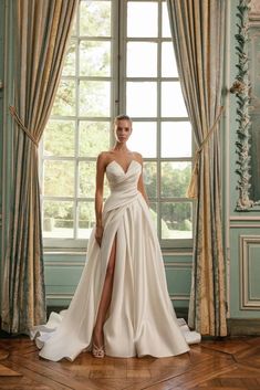 a woman standing in front of a window wearing a white dress with high slits