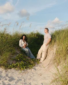 a man and woman sitting in the sand near tall grass