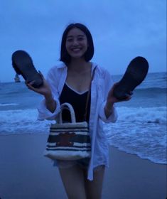 a woman standing on the beach holding up two shoes and a tote bag in front of her