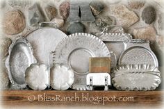 an assortment of silver plates and dishes on a shelf in front of a stone wall