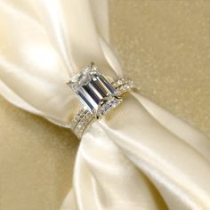 an emerald - cut diamond ring sits atop a white satin bow on a gold background