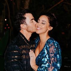 a man and woman kissing each other in front of some trees at night with lights behind them