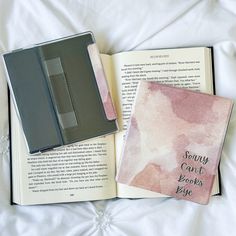 an open book sitting on top of a bed next to a pink and black notebook