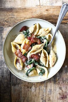 a bowl filled with pasta and spinach on top of a wooden table next to a spoon