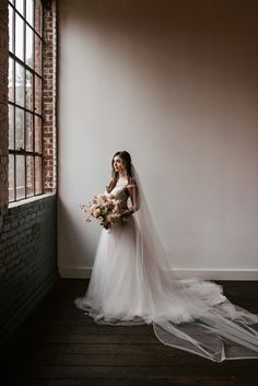 a woman standing in front of a window with a veil on her head and holding a bouquet