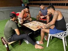 four men sitting around a table playing a board game on the grass in front of a building