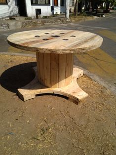 a round wooden table sitting on top of a dirt field next to a building and street