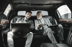 two men sitting in the back seat of a car looking at something on a laptop