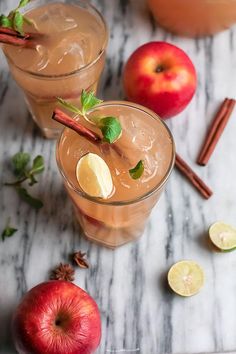 two glasses filled with apple cider on top of a marble table next to apples and cinnamon sticks