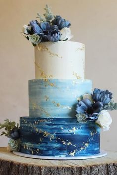 a three tiered blue and white cake with flowers on top is sitting on a tree stump