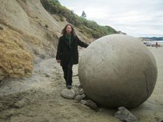 a woman standing next to a large stone ball on the side of a cliff covered in sand