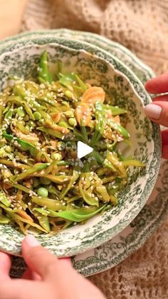 someone holding a bowl filled with green vegetables and sprouts on top of a table