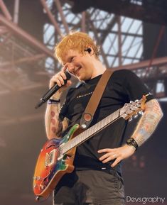 a man holding a guitar and singing into a microphone while standing in front of a stage