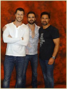 three men standing next to each other in front of a red wall with their arms crossed