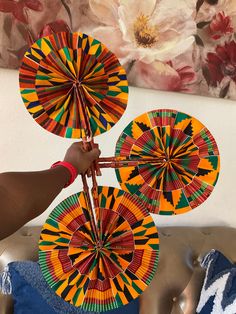 someone holding three colorful paper fans in front of a floral wall hanging on the wall