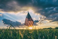 an american flag is in the grass under a cloudy sky with sun peeking through clouds