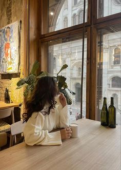 a woman sitting at a table in front of a window drinking from a coffee cup