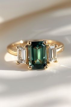 an emerald and diamond three stone ring on a white surface with shadows in the background