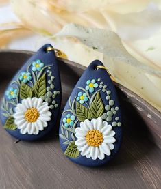 two blue earrings with white and yellow flowers are on a wooden tray next to a plant