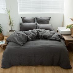 a bed with grey linens and pillows in a white room next to potted plants