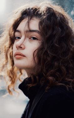 a close up of a person with curly hair looking off to the side and wearing a black jacket