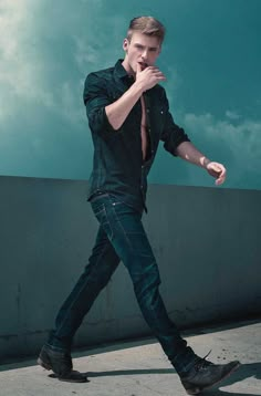 a man in black shirt and jeans walking on cement ground with his hand under his chin