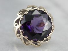 "A gorgeous purple amethyst with flashes of indigo and plenty of glitter is the centerpiece of this fabulous mid-century cocktail ring. A pierced scrolling scallop frame adds graphic interest and makes this piece unique to other ornate cocktail rings! Metal: 14K Yellow Gold Gem: Amethyst 17.69 Carats Gem Measurements: 17.2 mm, Round Ring Size: 6 Marks: \"14K\" Stamped on the inside band SKU #: N7UDRCEV Each piece has been identified and graded by a Graduate Gemologist who has been certified by t Formal Purple Sapphire Ring With Gemstone Accents, Purple Amethyst Art Deco Ring For Anniversary, Elegant Purple Sapphire Ring With Center Stone, Art Deco Purple Amethyst Ring For Anniversary, Victorian Style Purple Amethyst Ring For Anniversary, Luxury Purple Sapphire Ring For Formal Occasions, Purple Amethyst Jewelry For Evening, Purple Ring With Gemstone Accents For Formal Occasions, Formal Purple Amethyst Ring With Gemstone Accents