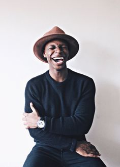 a man wearing a hat and black sweater sitting on a stool with his arms crossed