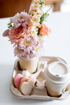 an arrangement of flowers in a vase on a tray next to cookies and a cup