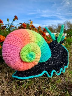 a crocheted snail is laying on the ground in front of some colorful flowers